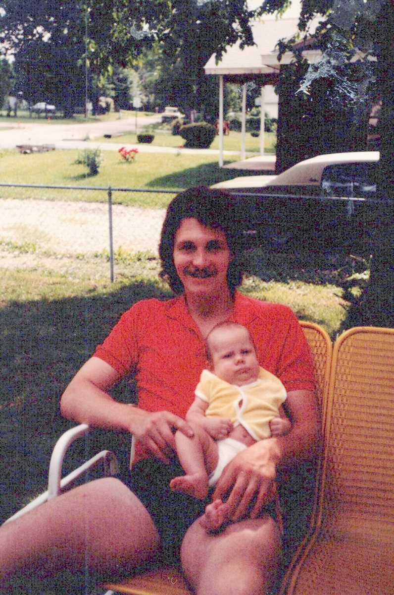 Mike Dixon in a red shirt, holding his infant son Shane on his lap outdoors